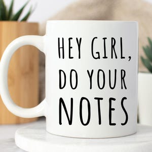May include: White ceramic mug with a curved handle. The mug features the text "HEY GIRL, DO YOUR NOTES" in black, handwritten-style lettering. The mug is sitting on a white, round coaster.