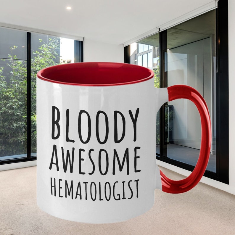 May include: White ceramic coffee mug with a red rim and handle. The mug has black text that reads "BLOODY AWESOME HEMATOLOGIST".