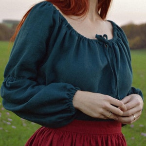 Op de afbeelding: Een vrouw draagt een tealblauwe boerenblouse met lange mouwen en een geplooide halslijn. Ze draagt ook een bordeauxrode rok.