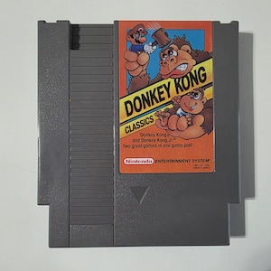 Puede incluir: Un cartucho gris de Nintendo Entertainment System (NES) con el juego "Donkey Kong Classics." La etiqueta muestra el t&iacute;tulo del juego, los personajes y el logotipo de Nintendo. El cartucho es rectangular con un borde biselado.