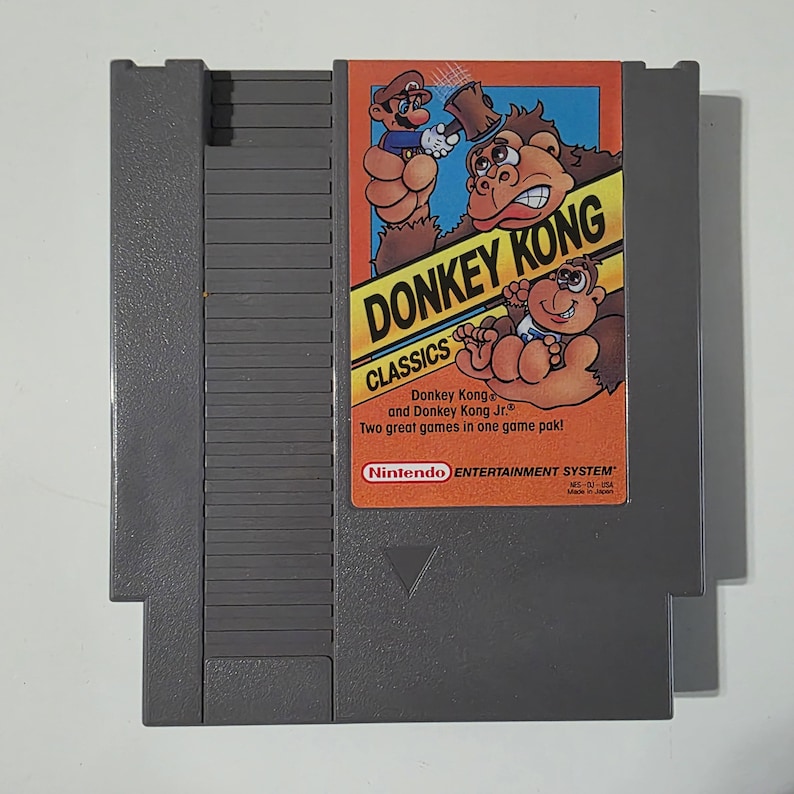 Puede incluir: Un cartucho gris de Nintendo Entertainment System (NES) con el juego "Donkey Kong Classics." La etiqueta muestra el t&iacute;tulo del juego, los personajes y el logotipo de Nintendo. El cartucho es rectangular con un borde biselado.
