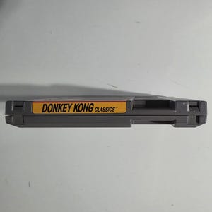 Puede incluir: Un cartucho de videojuego gris de Donkey Kong Classics. La etiqueta es amarilla y roja con el t&iacute;tulo del juego en texto negro. El cartucho es rectangular y tiene una ranura en el lateral.