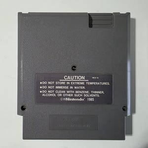 Puede incluir: Un cartucho de juego de Nintendo gris con el texto "CAUTION" e instrucciones de seguridad. El cartucho tiene forma rectangular con esquinas redondeadas y una peque&ntilde;a muesca en la parte superior. El logotipo de Nintendo y el a&ntilde;o 1985 tambi&eacute;n son visibles.