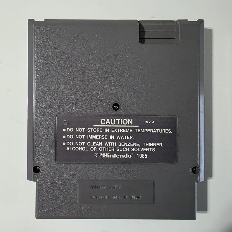 Puede incluir: Un cartucho de juego de Nintendo gris con el texto "CAUTION" e instrucciones de seguridad. El cartucho tiene forma rectangular con esquinas redondeadas y una peque&ntilde;a muesca en la parte superior. El logotipo de Nintendo y el a&ntilde;o 1985 tambi&eacute;n son visibles.
