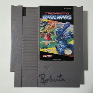 May include: A gray Nintendo Entertainment System (NES) cartridge for the game "Base Wars." The label features a futuristic baseball scene with robots. The cartridge has the "Ultra" and "Nintendo" logos, and the text "Made in Japan."