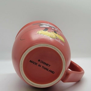 May include: A red ceramic mug with a Mickey Mouse design. The mug has a white rim and a white handle. The bottom of the mug reads "&copy; DISNEY MADE IN THAILAND".