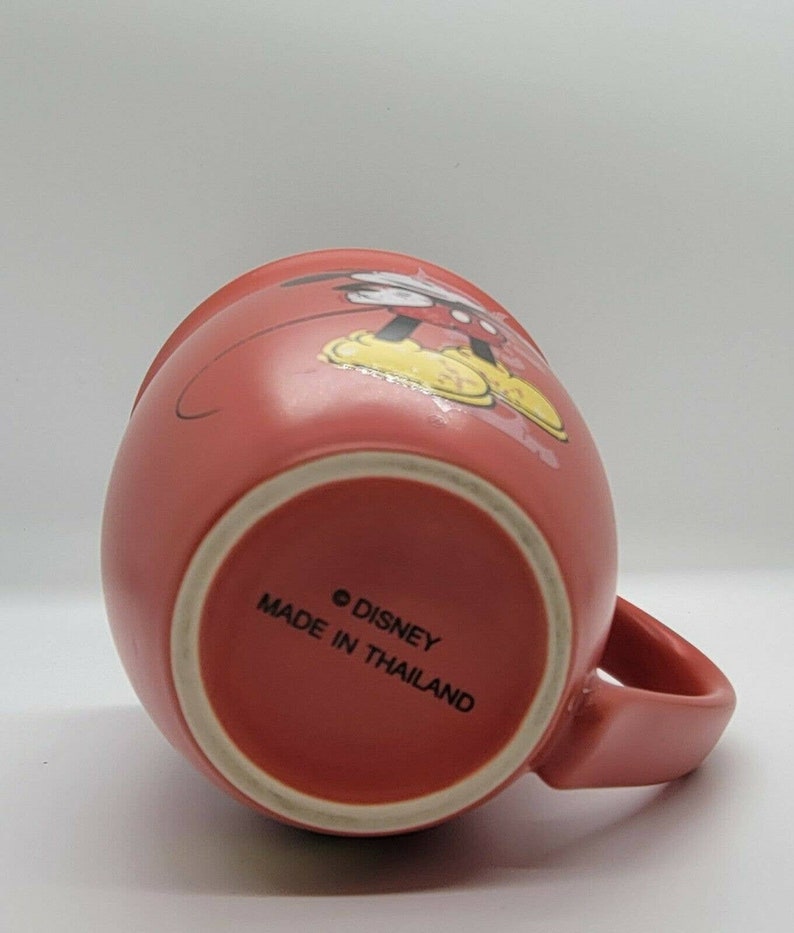 May include: A red ceramic mug with a Mickey Mouse design. The mug has a white rim and a white handle. The bottom of the mug reads "&copy; DISNEY MADE IN THAILAND".