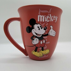 May include: Red ceramic coffee mug with a cartoon image of Mickey Mouse in a red and yellow outfit. The mug has the word "mickey" printed on it in white.