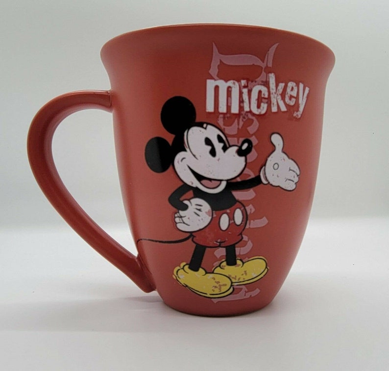 May include: Red ceramic coffee mug with a cartoon image of Mickey Mouse in a red and yellow outfit. The mug has the word "mickey" printed on it in white.