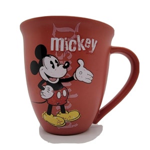 May include: Red ceramic coffee mug with a cartoon image of Mickey Mouse in a red and black outfit. The mug has the word "mickey" printed on it in white.