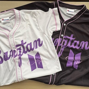 May include: Two short-sleeved button-down baseball jerseys, one white and one black, both featuring the purple text Bangtan and a purple logo with a heart outline. Each jersey has a V-neck collar with thin pink piping and a white inner neck label.