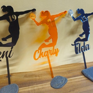 Könnte beinhalten: Drei schwarze, orange und blaue Acrylsilhouetten von weiblichen Sportlerinnen, die Volleyball spielen. Jede Silhouette trägt einen Namen und eine Nummer. Die Namen sind "Teni", "Charly" und "Tilda". Die Nummern sind 19, 1 und 13. Jede Silhouette ist auf einer schwarzen Metallstange und einem grauen Steinsockel montiert.