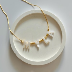 May include: A gold chain necklace spelling "MILA" in white letter beads. The necklace is presented on a white, round, textured surface. Gold-coloured beads are between the letters.