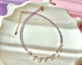 May include: A delicate purple beaded necklace with a gold clasp and white shell letter charms spelling out the name "SOPHIA".
