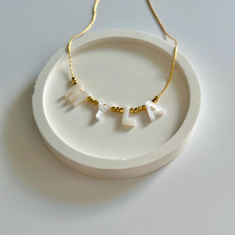 May include: A gold chain necklace spelling "Myla" in white letter beads. The necklace is presented on a white, circular, shallow dish. Gold-coloured beads are between the letters.
