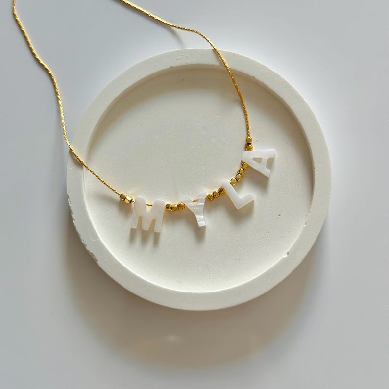 May include: A gold chain necklace spelling "MILA" in white letter beads. The necklace is presented on a white, round, textured surface. Gold-coloured beads are between the letters.