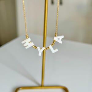 May include: A gold chain necklace featuring the letters "MYLA" in white, block-style letters. The letters are connected by small gold beads. The necklace is presented on a gold stand.