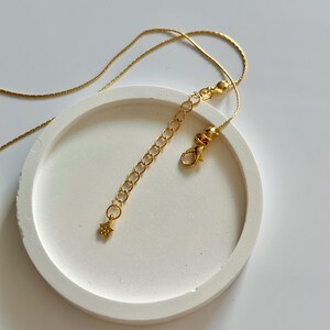 May include: A gold-coloured necklace with a star charm and an adjustable chain. The necklace is presented on a white, circular surface. The clasp and chain extender are visible, highlighting the necklace's design.