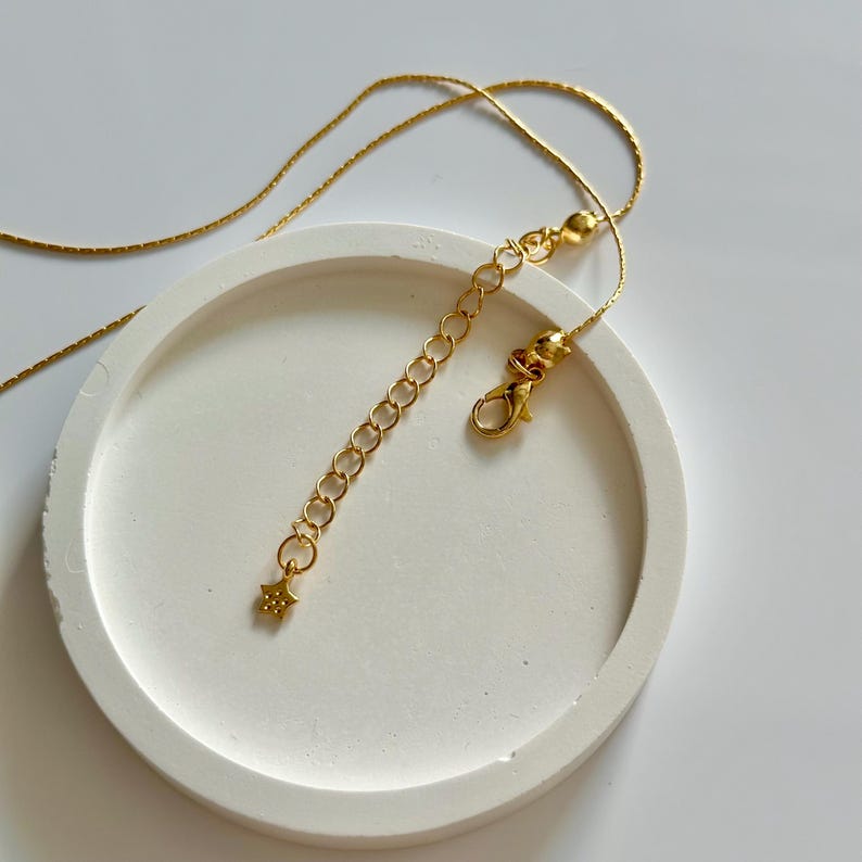 May include: A gold-coloured necklace with a star charm and an adjustable chain. The necklace is presented on a white, circular surface. The clasp and chain extender are visible, highlighting the necklace's design.