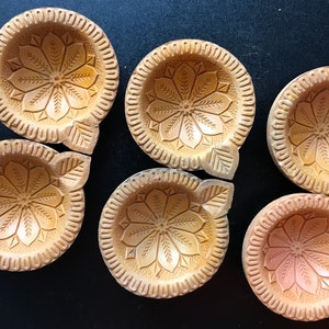 Op de afbeelding: Zes beige, gebeeldhouwde, cirkelvormige kaarsenhouders met bloemen- en bladerenontwerpen. Elke houder heeft een centraal bloemmotief omgeven door een decoratieve rand. De houders zijn gerangschikt op een donkere ondergrond.