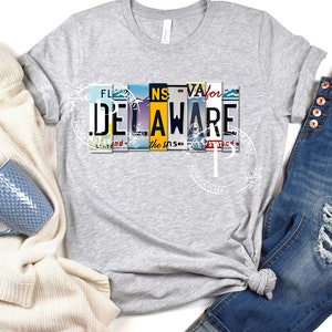 Peut inclure: Un t-shirt gris avec un motif de plaques d'immatriculation qui épellent "DELAWARE". Les plaques d'immatriculation proviennent de différents États, dont la Floride, la Virginie et la Caroline du Nord.