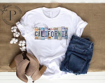 Puede incluir: Camiseta blanca con un gráfico de la palabra "CALIFORNIA" hecha de placas de matrícula antiguas. El gráfico está en tonos de azul, verde y rojo. La camiseta se muestra sobre un fondo de arpillera marrón con un par de botines marrones y unos vaqueros azules.