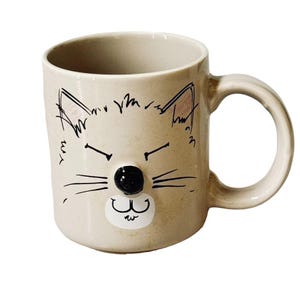 May include: Beige ceramic mug with a whimsical cat face design. The cat face features black drawn whiskers, eyebrows, and ear outlines, with a black 3D nose and a white mouth. The mug has a rounded handle.