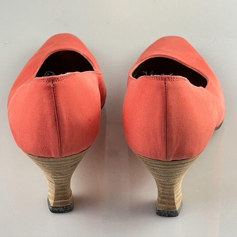 May include: A pair of coral colored high heeled shoes with a low heel. The shoes are made of a soft fabric and have a pointed toe.