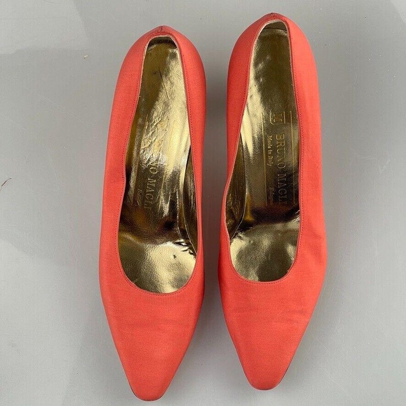 May include: A pair of coral-colored satin pumps with a pointed toe and a low heel. The shoes are made by Bruno Magli and are made in Italy.
