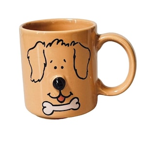 May include: A light tan ceramic mug featuring a cartoon dog face illustration. The design includes a raised, three-dimensional black nose, a small pink tongue, and a white bone graphic positioned below the mouth. The mug has a curved handle.