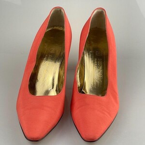 May include: A pair of coral colored high-heeled pumps with a pointed toe. The shoes are made of a satin-like fabric and have a gold leather insole with the text "Bruno Magli Made in Italy" printed on it.