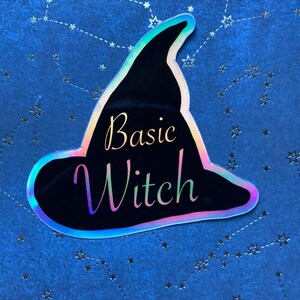 May include: A black witch hat sticker with iridescent edging and the words "Basic Witch" in a script font. The sticker is set against a blue background with a constellation pattern.