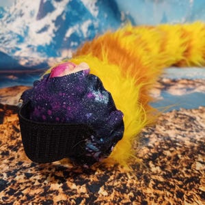 May include: A plush, cat-like tail with a vibrant color scheme. The tail features a fluffy, yellow and orange striped design. The base is a dark fabric with a galaxy print, and a black elastic band.