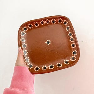 May include: A square, brown ceramic tray with a decorative border of white and black floral designs. The tray has a central floral design and is held against a white background.