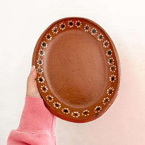 May include: Oval terracotta plate with a brown center and a decorative border of small white and black floral designs. The plate is held against a white background, showcasing its rustic charm and handcrafted aesthetic.