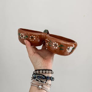 May include: Handmade brown ceramic serving dish with two connected bowls. Each bowl is decorated with white and green floral patterns. The dish is held up against a white background.