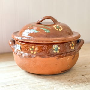 May include: A round brown clay pot with a matching lid and two side handles. The exterior features a glossy finish with hand-painted yellow and green floral motifs. A small white rectangular sticker is visible on the left side of the pot.