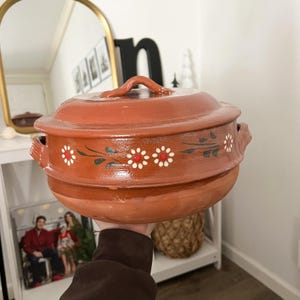May include: A terracotta-colored ceramic casserole dish with a lid and handles. The dish is decorated with white daisy-like flowers and green leaves. The lid has a handle for easy lifting.