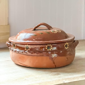 May include: A round terracotta casserole dish with a matching lid, featuring a glossy brown glaze on the upper half and a matte natural clay finish on the bottom. The exterior is decorated with hand-painted yellow and green floral motifs.