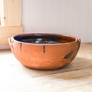 May include: A round terracotta bowl with two small side handles. The exterior has a natural matte clay finish with dark brown glaze drips, while the interior features a smooth, dark brown glazed surface. The bowl has a wide, open top.