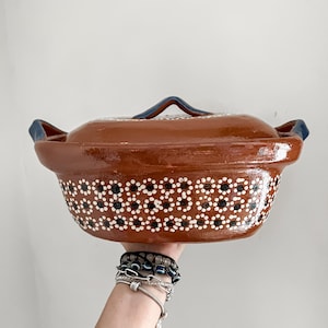 May include: A brown ceramic casserole dish with a lid. The dish features a decorative pattern of white and black dots around the rim. The lid has a blue handle. The dish is held up by a person's hand.