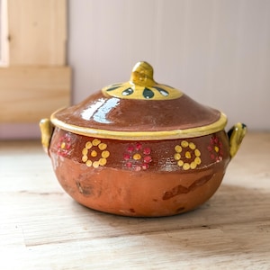 May include: A round, brown clay pot with a matching lid, featuring two side handles. The upper portion of the pot and the lid are decorated with hand-painted yellow and red floral patterns. The surface has a glossy, glazed finish throughout.