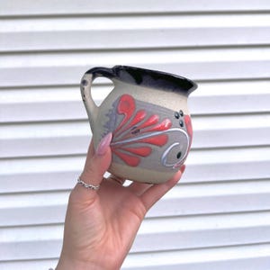 May include: A small, handcrafted ceramic mug with a handle. The mug features a beige base with red and gray floral designs and a dark blue rim. The mug is held in a hand.