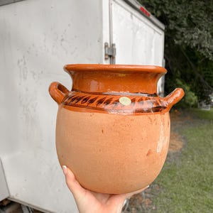 May include: A terracotta-colored, round clay pot with two handles. The pot has a wide, flared rim and a band of decorative brown and black leaf-like designs around the middle. The pot is held in a hand.