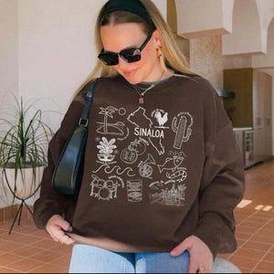 May include: A brown sweatshirt with a white graphic design featuring the word "SINALOA" and various illustrations. The design includes a map, a rooster, a cactus, and other symbols. The sweatshirt is worn by a person wearing sunglasses.