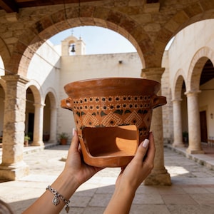 May include: A handmade, terracotta clay brazier with a decorative pattern of black and white dots and green leaf-like shapes. The brazier has two handles and a rectangular opening. The background features an arched stone structure.