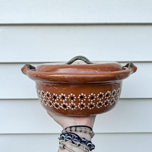 May include: A handmade, terracotta-colored ceramic casserole dish with a matching lid. The dish features a floral pattern of white dots with black centers. The lid has a green handle. The dish is held up against a white background.