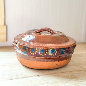 May include: A round, brown clay pot with a matching lid, featuring a glossy finish and a hand-painted decorative band around the center. The design includes blue floral motifs surrounded by small yellow dots on a dark brown background.