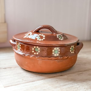 May include: A round terracotta cooking pot with a matching lid, featuring two side handles and a decorative band of painted white and green flower motifs. The clay surface has a natural reddish-brown tone with a smooth, glazed finish on the upper section.