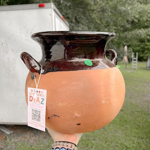 May include: A handmade, terracotta-colored ceramic pot with a dark brown glazed rim and handles. The pot has a rounded body and a tag that reads "ART SANIA DIAZ". The pot is held up by a person.